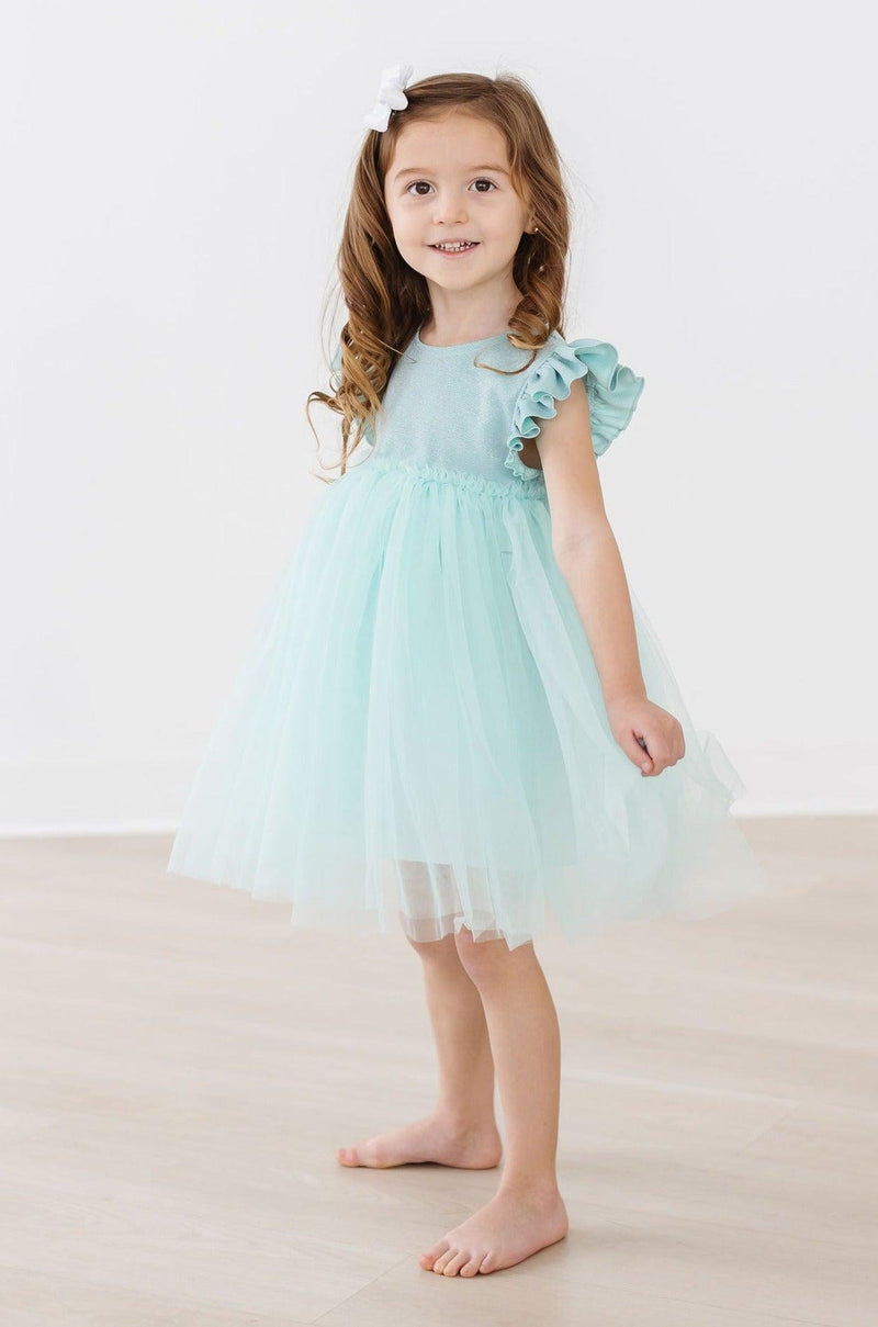 Sage Shimmer Tutu Dress displayed on a cute girl standing gracefully, twirling her sage green tutu dress with ruffled sleeves for a playful look.