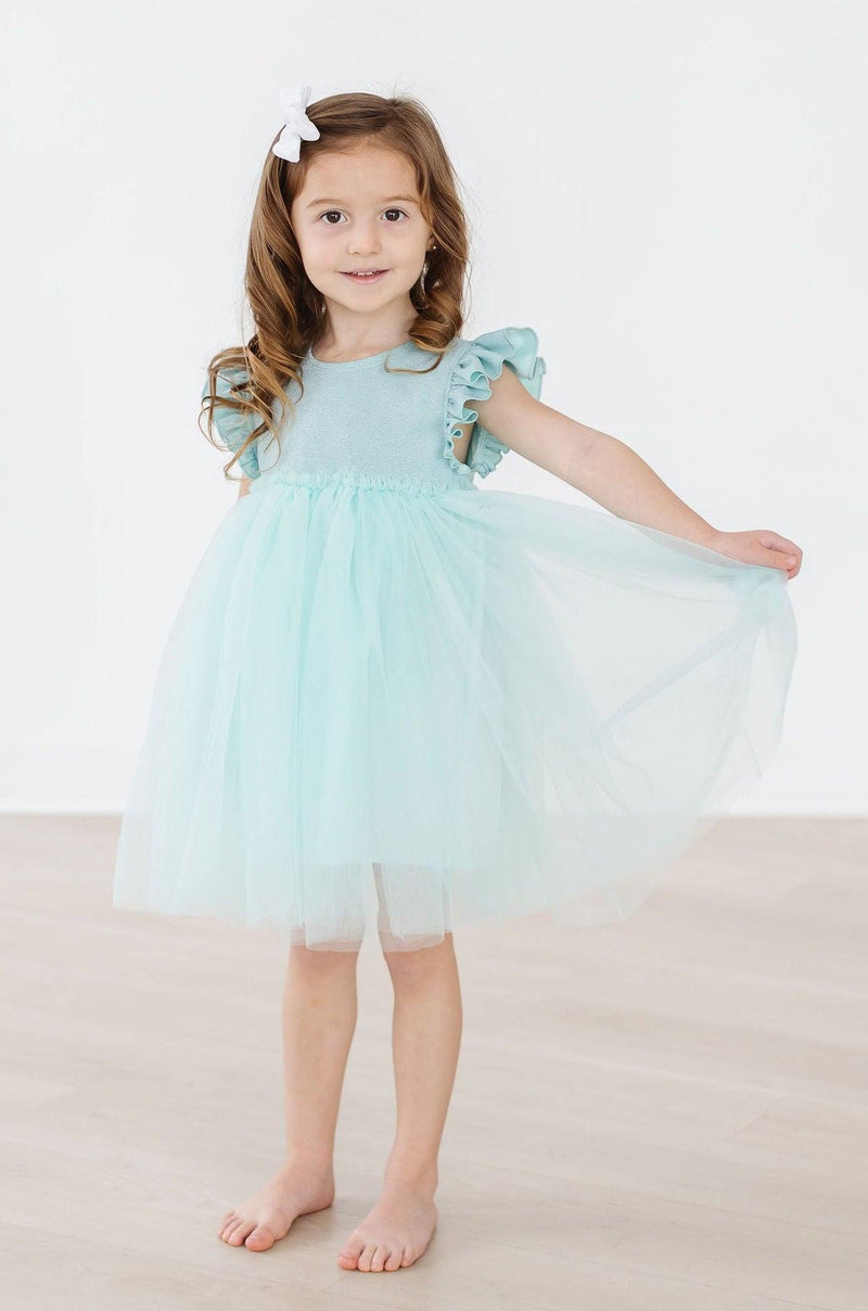 Sage Shimmer Tutu Dress on a young girl with curly hair, wearing a sage green tutu dress with flutter sleeves, showcasing a joyful pose.