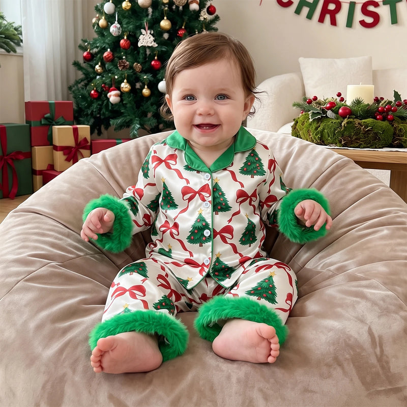 Baby wearing Christmas tree and red bow print pajama set with green puffy cuffs, sitting on beanbag near decorated tree.