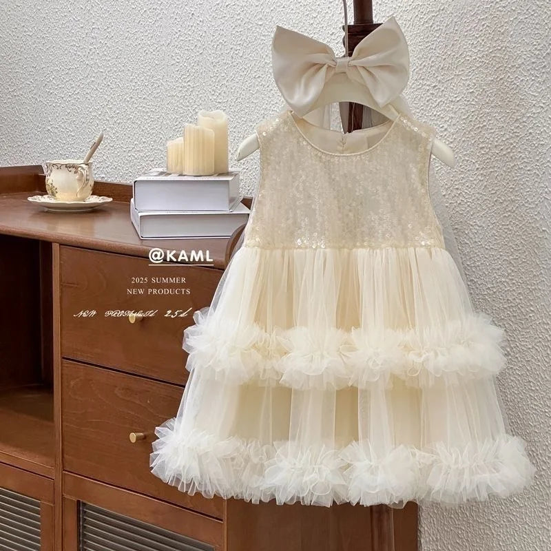 A cream-colored toddler girl's party dress with a large bow headband, sequined bodice, and tiered tulle skirt, displayed on a wooden hanger next to a wooden cabinet.
