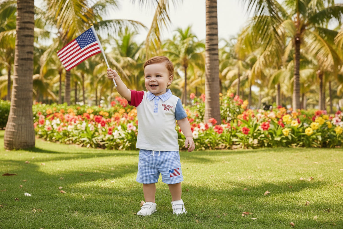 Toddler boy in American Boy patriotic polo shirt and blue seersucker shorts, holding an American flag in a sunny park.