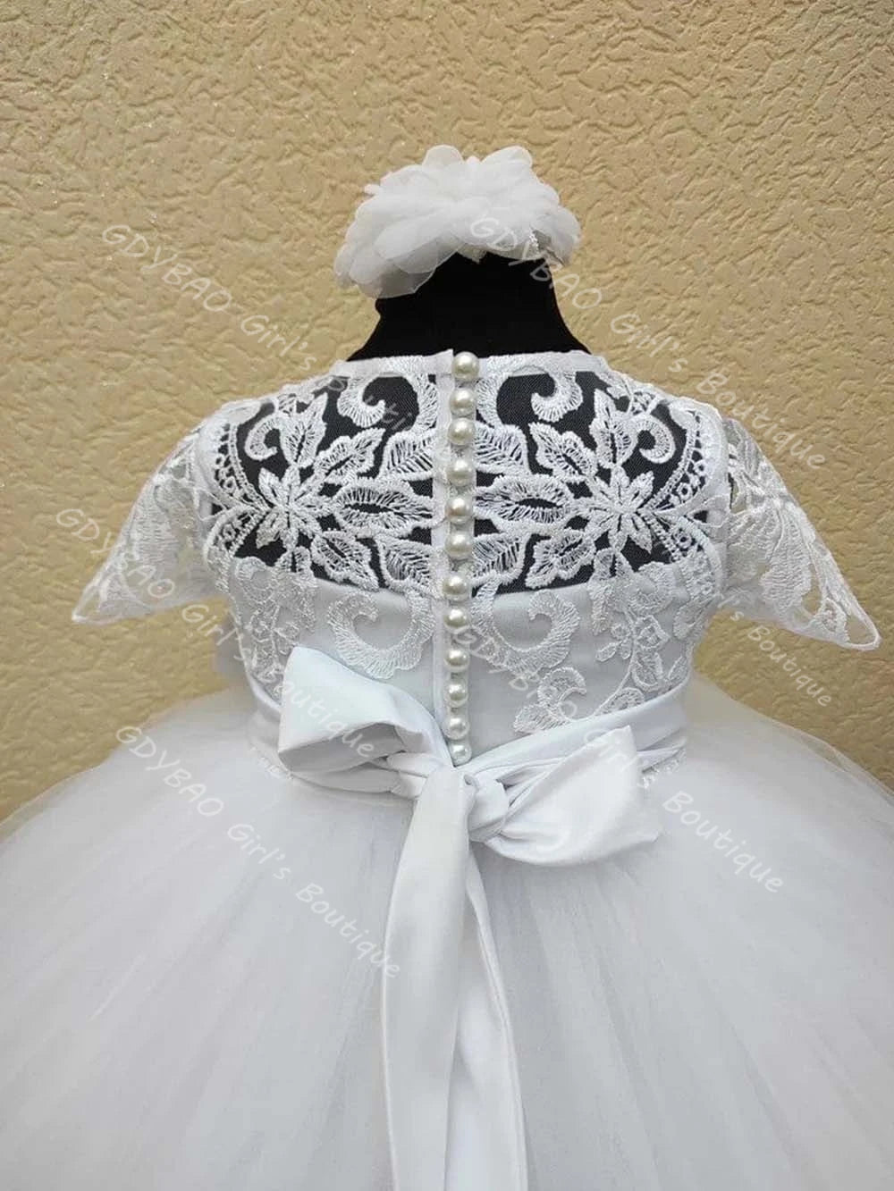 White lace flower girl dress back view featuring beaded pearl buttons, floral applique, and large satin bow sash for wedding or special occasion.