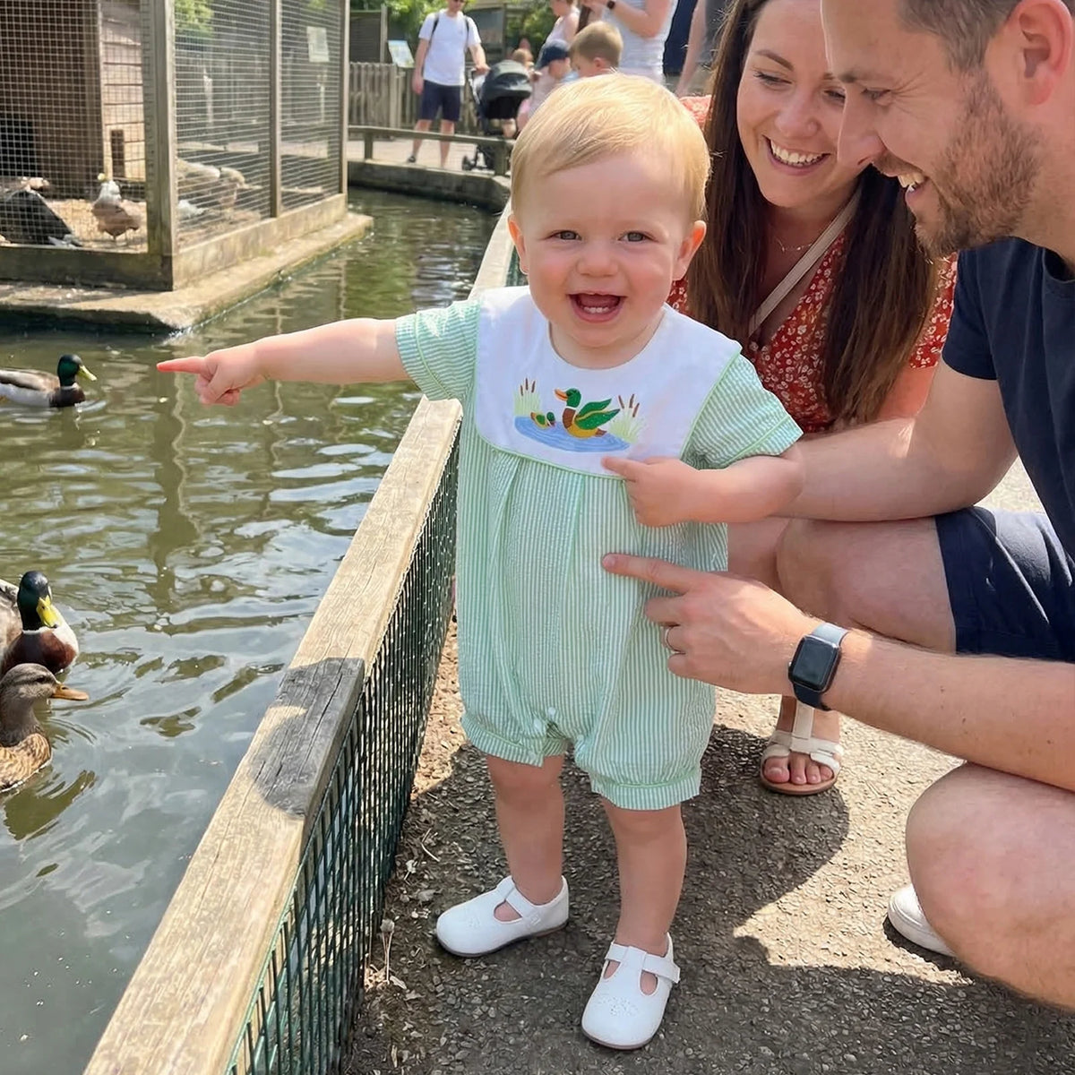 Baby boy in green striped bibbed seersucker romper with a duck-embroidered bib beside a pond.