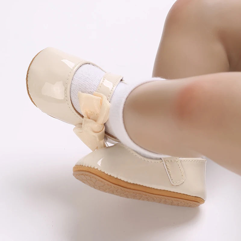 Infant wearing 'Serena' Bow First Walker Shoes in glossy beige with a butterfly-knot bow, designed for style and comfort.