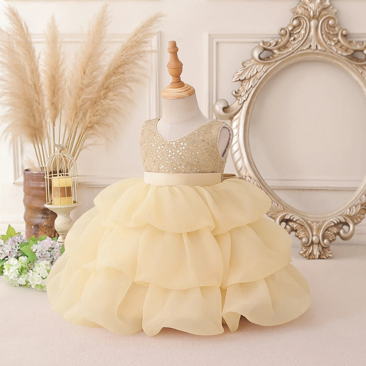 A toddler girl's formal party dress displayed on a mannequin, featuring a sparkling champagne sequined bodice and a voluminous, tiered cream tulle skirt. The dress is styled in a vintage-inspired room setting with a large ornate mirror, pampas grass, and small flowers.