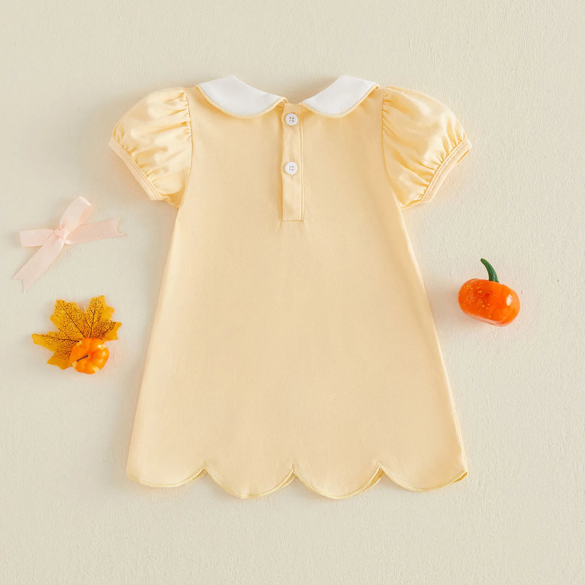 A pale yellow baby dress with a white Peter Pan collar and short puff sleeves, laid flat on a light beige background with decorative autumn props.