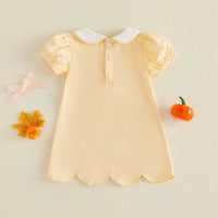 A pale yellow baby dress with a white Peter Pan collar and short puff sleeves, laid flat on a light beige background with decorative autumn props.