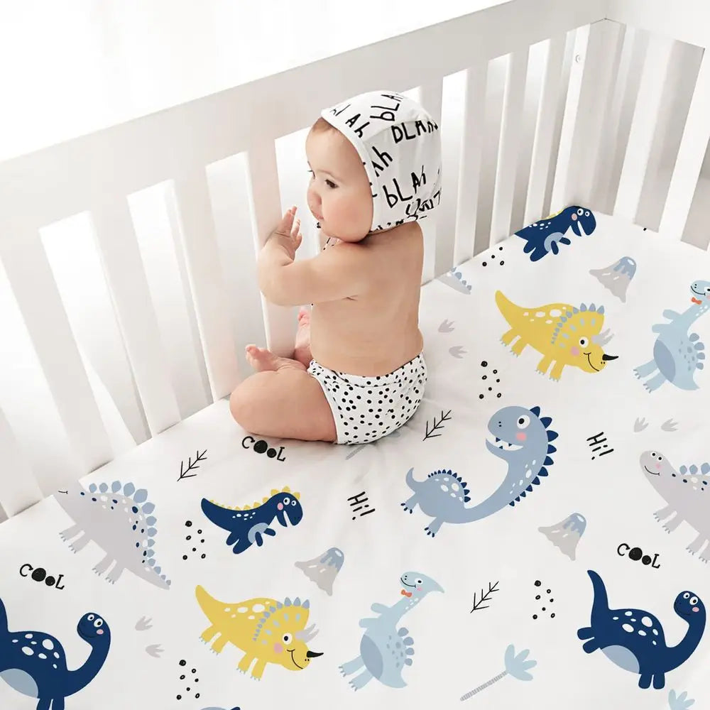 Baby sits in a white crib on dinosaur-print microfiber crib sheets (52×28 in), part of a 4-pack.