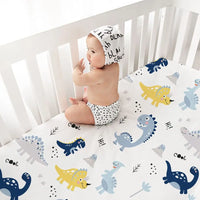 Baby sits in a white crib on dinosaur-print microfiber crib sheets (52×28 in), part of a 4-pack.