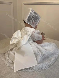 White lace baby girl baptism dress with sequins, ruffles, and large satin bow back, paired with a matching lace bonnet.