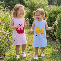 Two girls wear back-to-school seersucker dresses, sleeveless with embroidered bows (pink and blue) in a garden.
