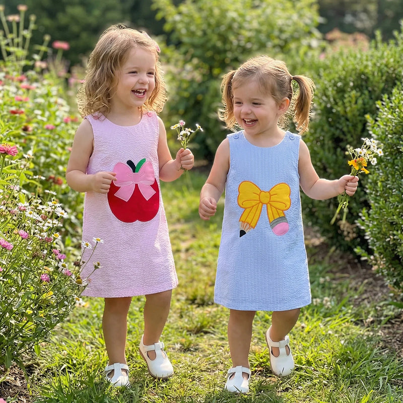 Two girls wear back-to-school seersucker dresses, sleeveless with embroidered bows (pink and blue) in a garden.