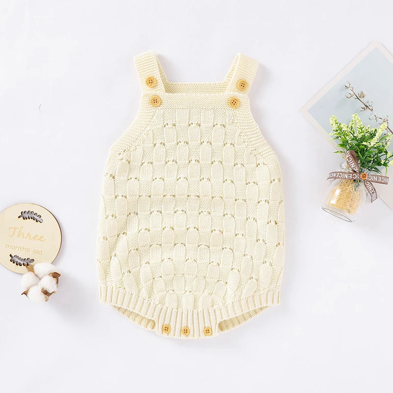 A cream-colored knit baby romper with shoulder straps and decorative wooden buttons, styled in a flat-lay arrangement with decorative props on a white background.