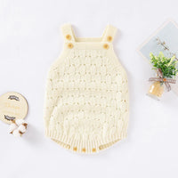 A cream-colored knit baby romper with shoulder straps and decorative wooden buttons, styled in a flat-lay arrangement with decorative props on a white background.