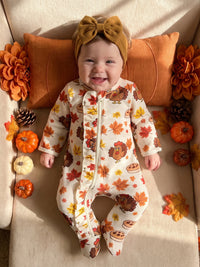 Baby wearing turkey and leaf print footed romper with ruffle front and mustard headbow, surrounded by pumpkins and fall decor.