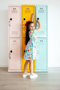 School Rules Short Sleeve Pocket Twirl Dress worn by a girl beside pastel lockers.