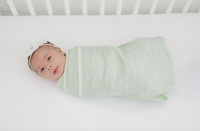 Newborn baby swaddled in GOTS certified organic cotton blanket, desert green with "I Love You" script pattern on white crib sheet.
