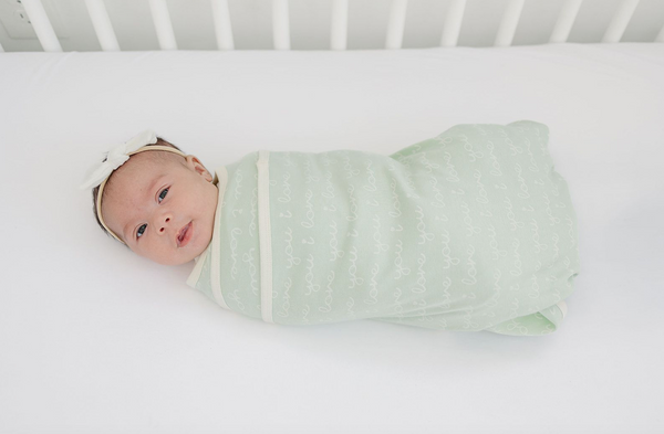 Newborn baby swaddled in GOTS certified organic cotton blanket, desert green with "I Love You" script pattern on white crib sheet.