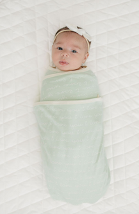 Baby swaddled in GOTS certified organic cotton desert green blanket with "I love you" print, lying on white quilted mat.