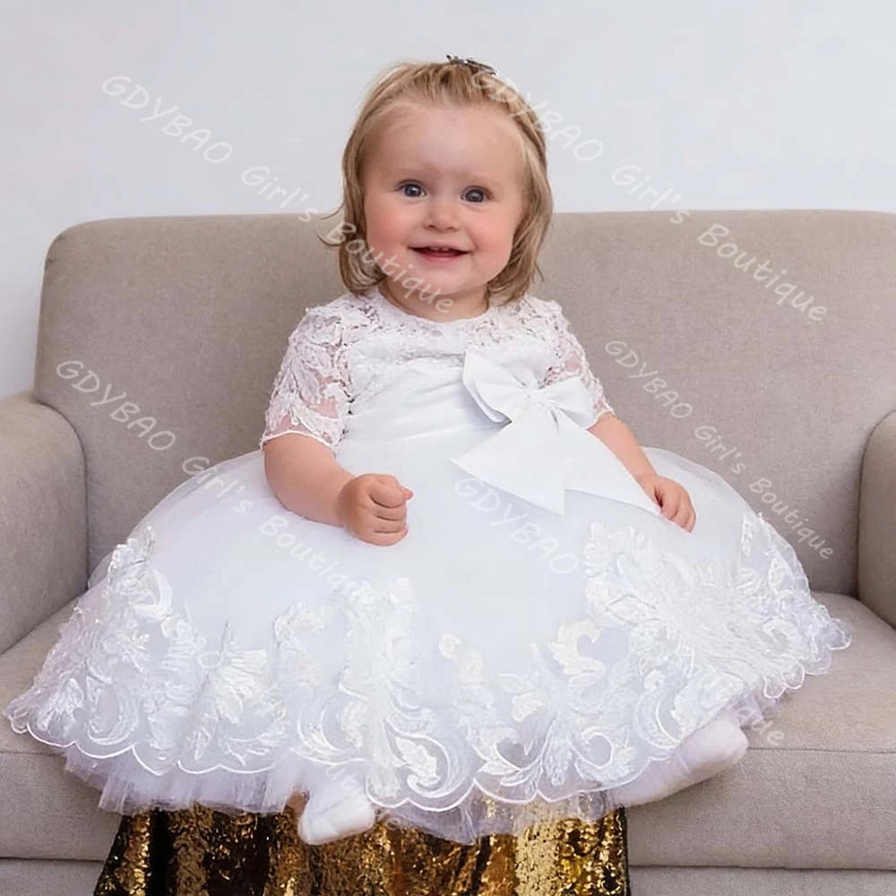 Toddler girl wearing a white lace special occasion dress with appliques, ruffles, and a large front bow for weddings or parties.