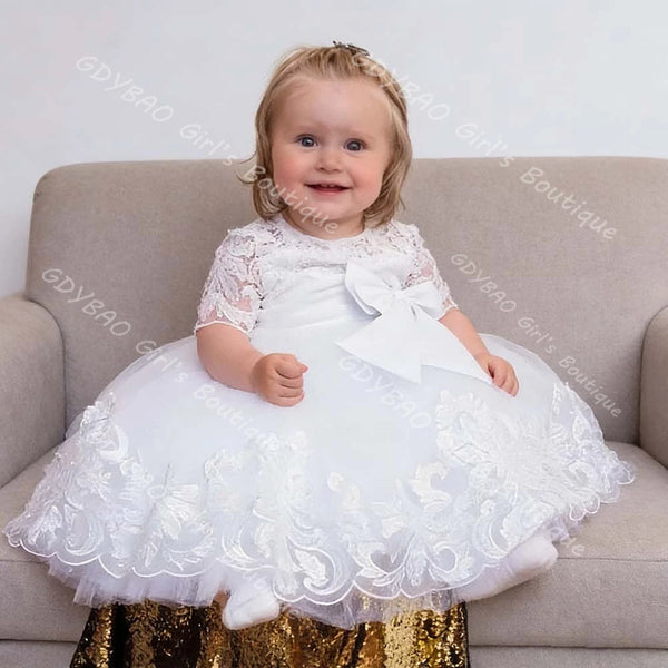 Toddler girl wearing a white lace special occasion dress with appliques, ruffles, and a large front bow for weddings or parties.