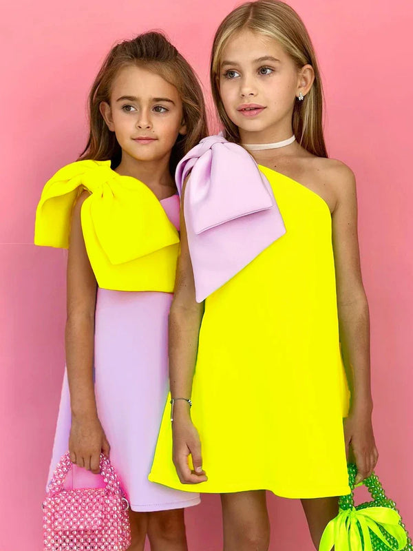 Two girls wearing the 'Logan' Chic Kids Party Dress, an off-shoulder yellow dress with a purple bow, showcasing style and fun, perfect for special occasions. Ideal chic kids party dress.