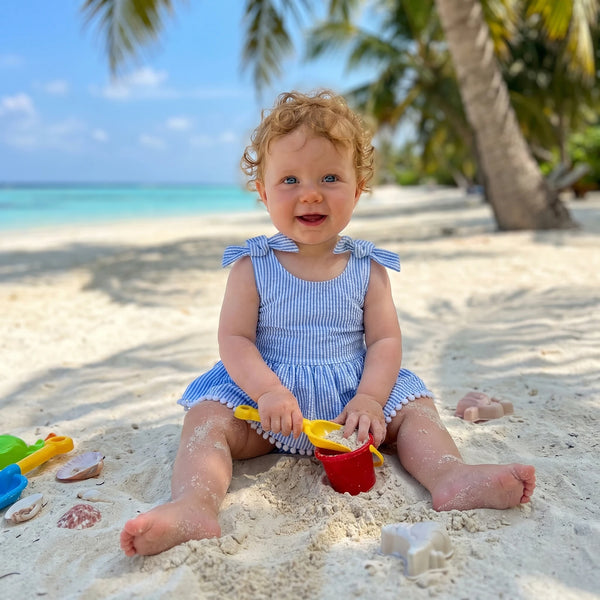 Baby wearing Suzie Seersucker Stripe Frilly Romper at the beach, blue-and-white stripes with shoulder bows.