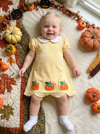 A baby lying on a quilted blanket surrounded by pumpkins, corn, and autumn decorations, wearing a pale yellow short-sleeve dress with a white Peter Pan collar and three orange pumpkin appliques near the hem.