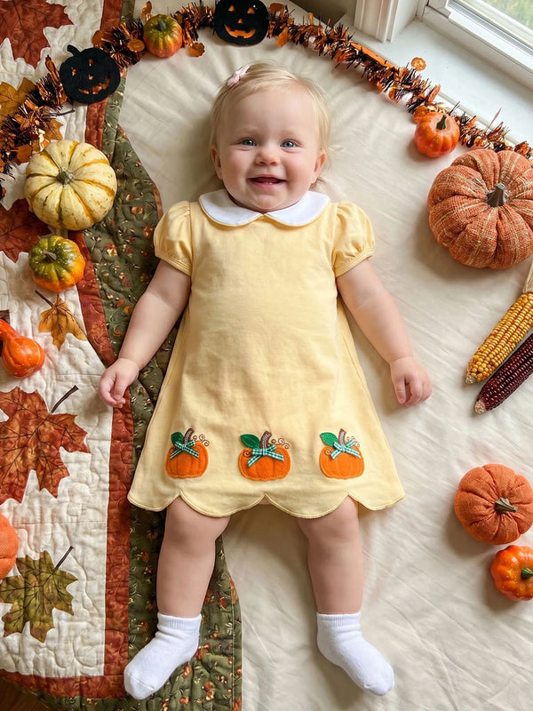 A baby lying on a quilted blanket surrounded by pumpkins, corn, and autumn decorations, wearing a pale yellow short-sleeve dress with a white Peter Pan collar and three orange pumpkin appliques near the hem.
