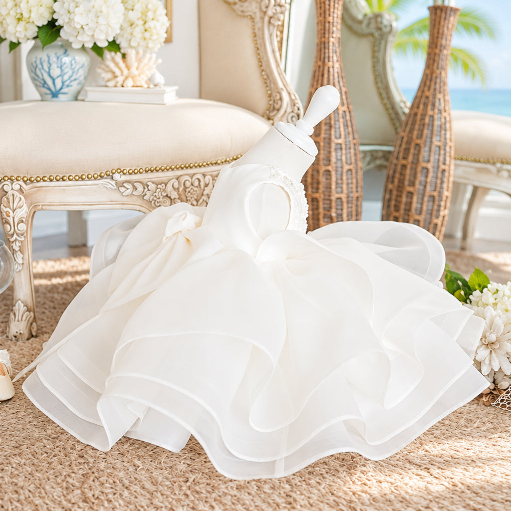 A white organza baby dress with a ruffled tulle skirt and a large bow, displayed on a white mannequin in an ornate vintage room.