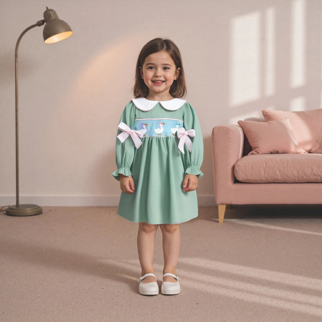 A young girl wearing the "Goosey Goose" Goose Embroidered Mint Smocked Long-Sleeve Dress, featuring a Peter Pan collar and charming embroidered geese, standing in a cozy room with a warm ambiance.