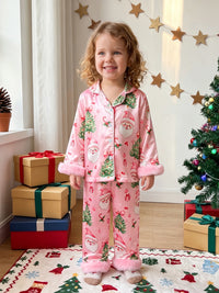 Girl wearing pink satin Christmas pajama set with Santa print and fuzzy cuffs, standing by gifts and a tree.