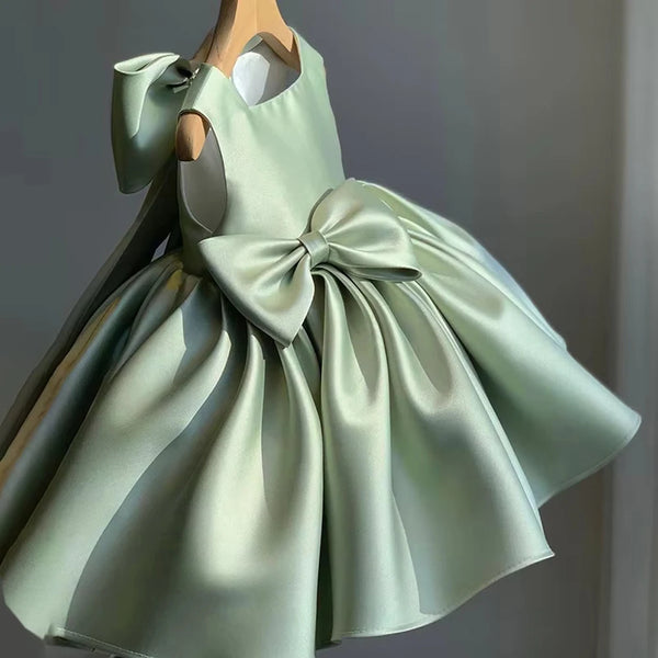 A pale mint green satin dress displayed on a wooden hanger against a neutral gray background. The dress features a halter-style neckline with a large bow on the shoulder, a matching waist bow, and a voluminous flared skirt.