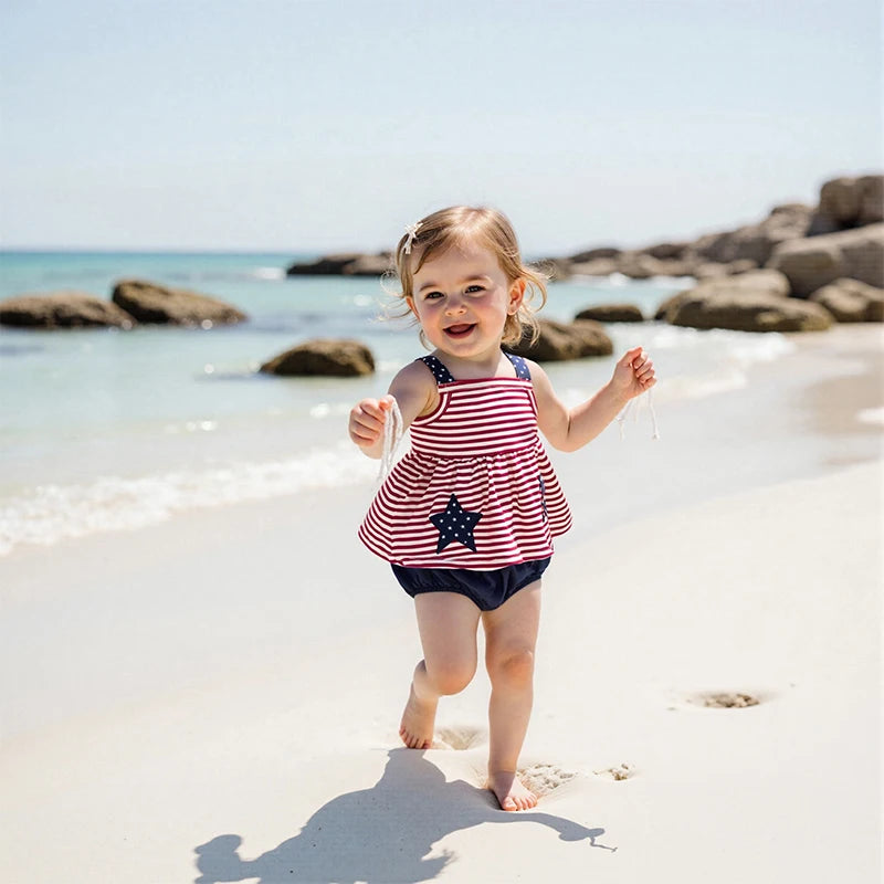 A playful moment of a girl at the beach in the 'Stars and Stripes' 2 PC Dress and Bloomer Set, with a focus on the colorful design and her beaming smile, ideal for summer outings.