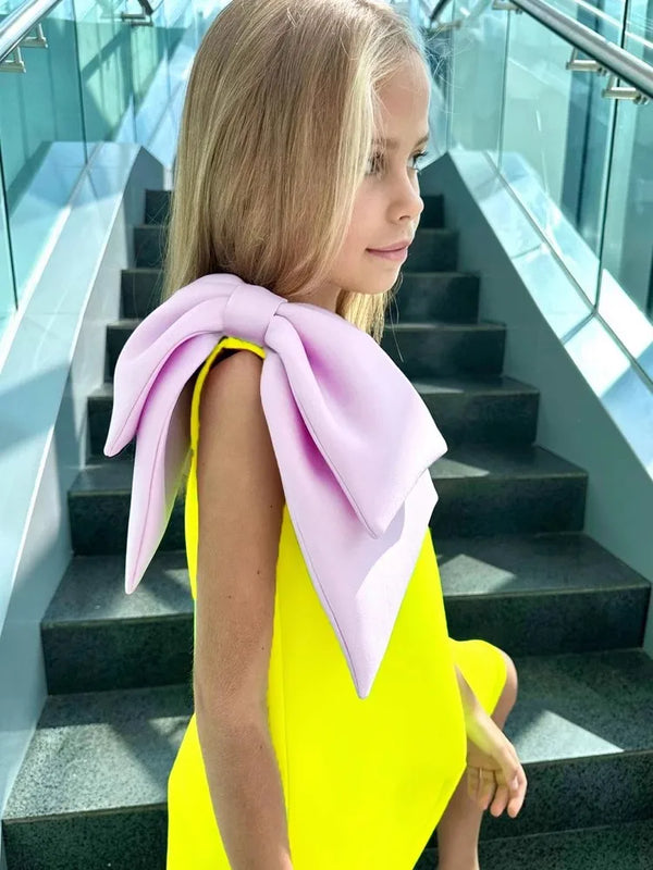 A close-up of a girl in the 'Logan' Chic Kids Party Dress, featuring a bright yellow color and a stunning purple bow on one shoulder, highlighting this chic kids party dress.