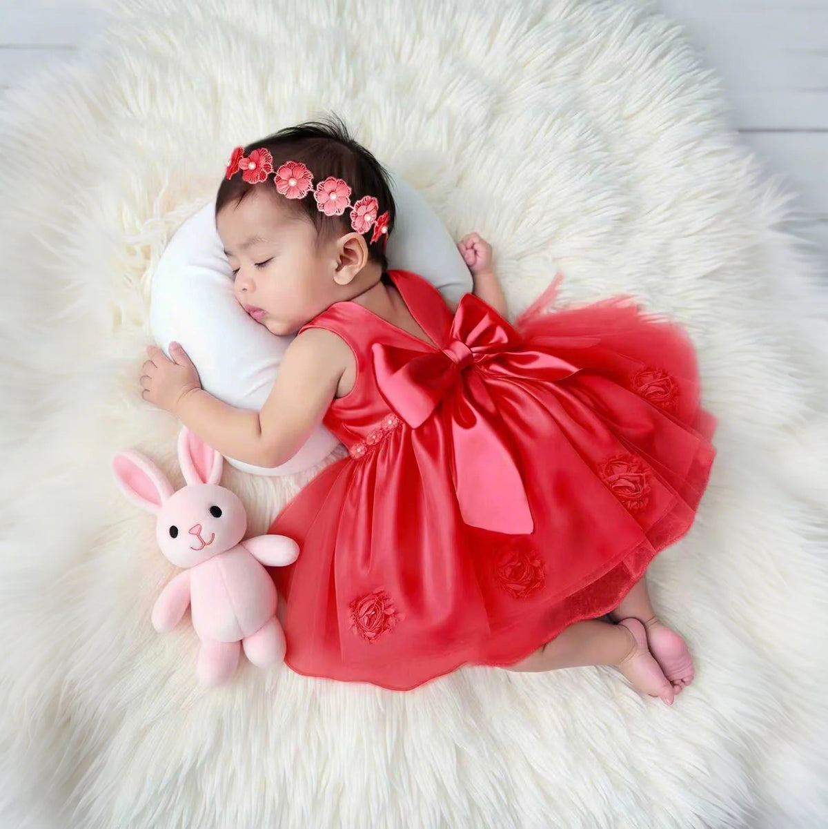 Sleeping baby in Lara Elegant infant dress in red with oversized bow and beaded headband on white fur rug.