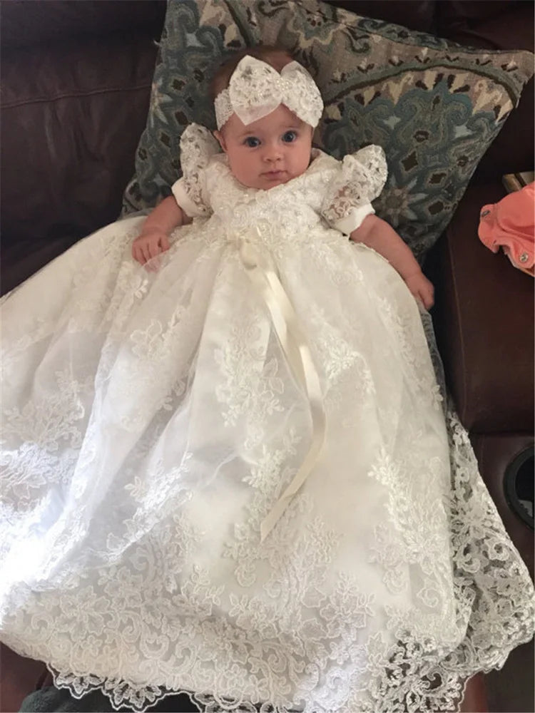 Baby girl wearing the 'Leonella' Elegant Lace Baptism Gown in white, featuring delicate lace and a satin ribbon, perfect for special ceremonies.