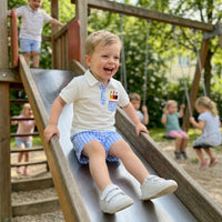 Boy wearing a white polo with pencil embroidery and blue check shorts, smiling on a playground slide.