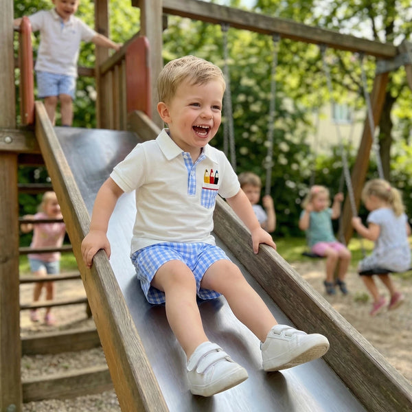 Boy wearing a white polo with pencil embroidery and blue check shorts, smiling on a playground slide.
