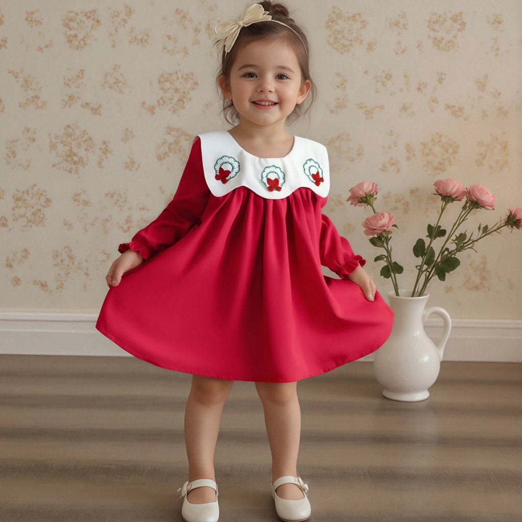 Adorable toddler girl wearing the Three Little Wreaths Bibbed Baby A-Line Dress in red, featuring a white bibbed collar with embroidered wreaths, perfect for holiday celebrations. She poses cutely with flowers in a cozy indoor setting.