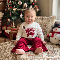 Toddler girl in white Holly Jolly pullover and red velvet bell bottom pants sitting by a Christmas tree.