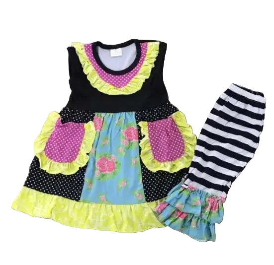 A toddler girl's two-piece patchwork outfit laid flat on a dark wood floor. The sleeveless top is black with a light blue floral center panel, pink polka-dot pockets with yellow ruffles, and a yellow ruffled hem.