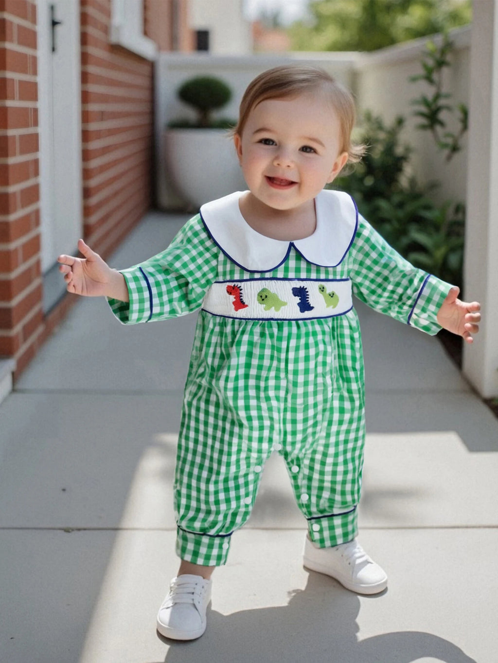 Adorable baby wearing the 'Dino-Might!' Long Sleeve Peter Pan Collar Check Romper, featuring a green and white checked pattern and smocked dinosaurs, standing on a patio.