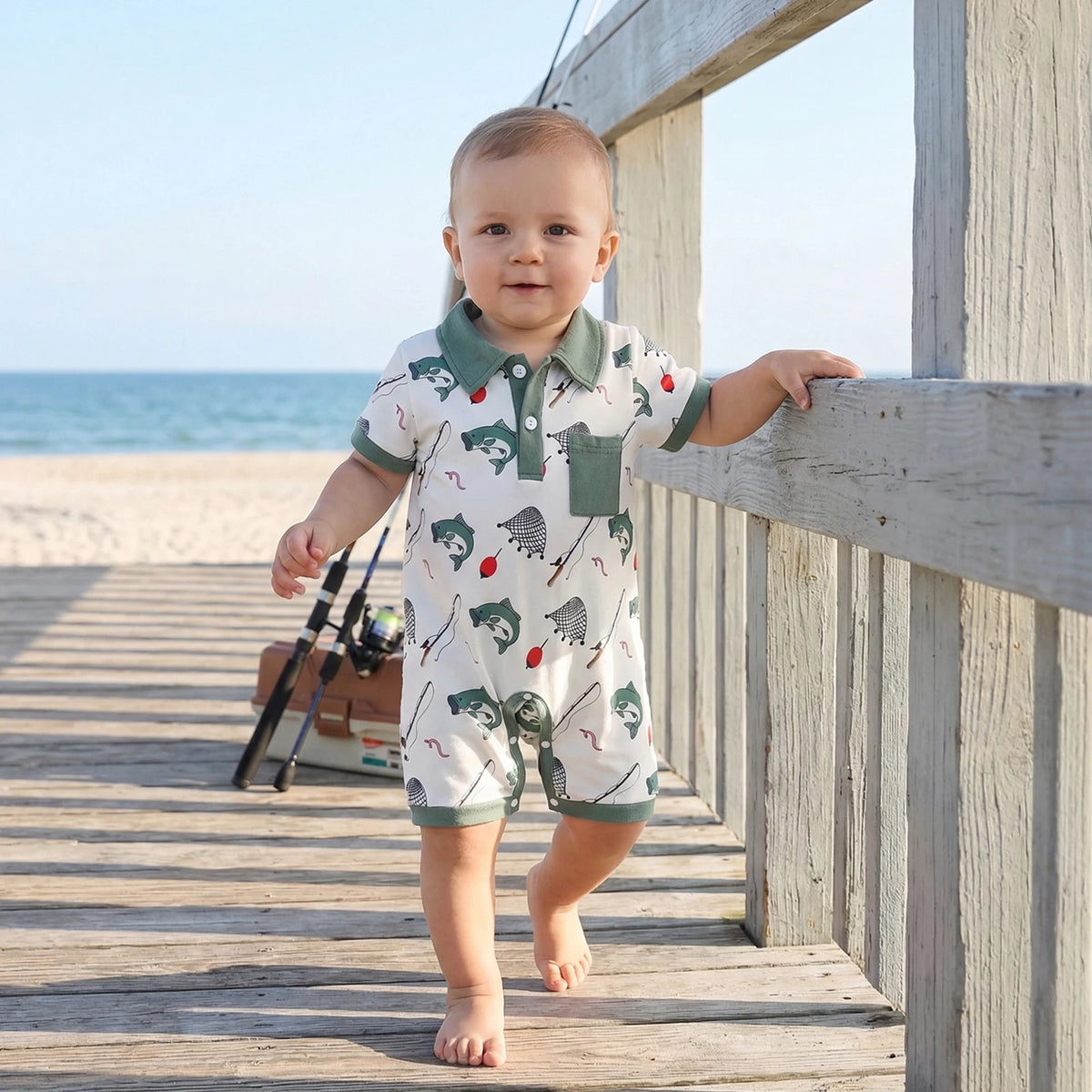 Baby boy in polo-style short-sleeve romper with fishing-themed print, green collar and pocket, at the beach boardwalk.