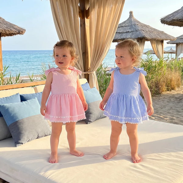 Two toddlers wearing Suzie seersucker stripe frilly romper swimsuits with tassel trim and shoulder bows at the beach.