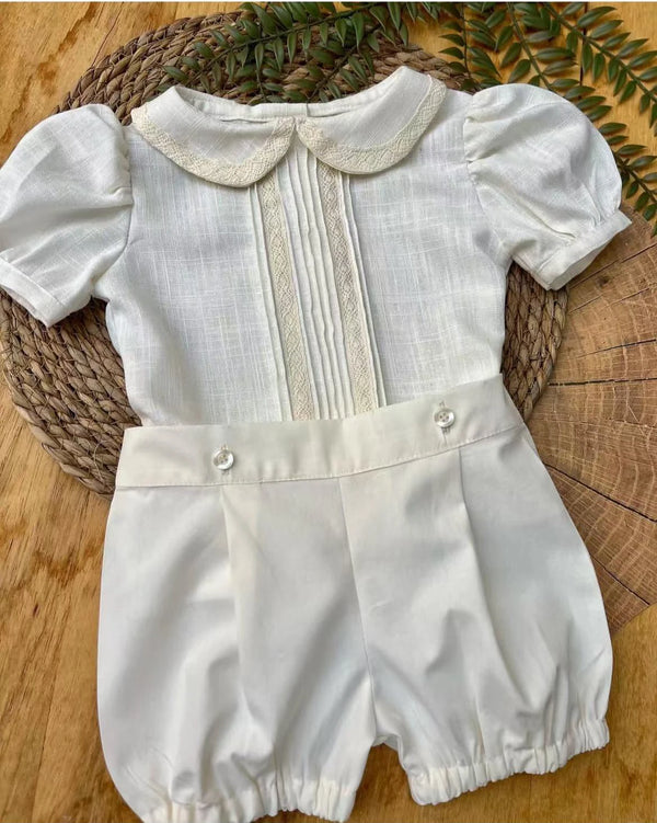 A baby boy's short-sleeve romper in off-white linen, featuring a Peter Pan collar with embroidered trim, vertical lace plissé pleats on the chest, and buttoned waist details, displayed on a woven mat over a wood slice background.