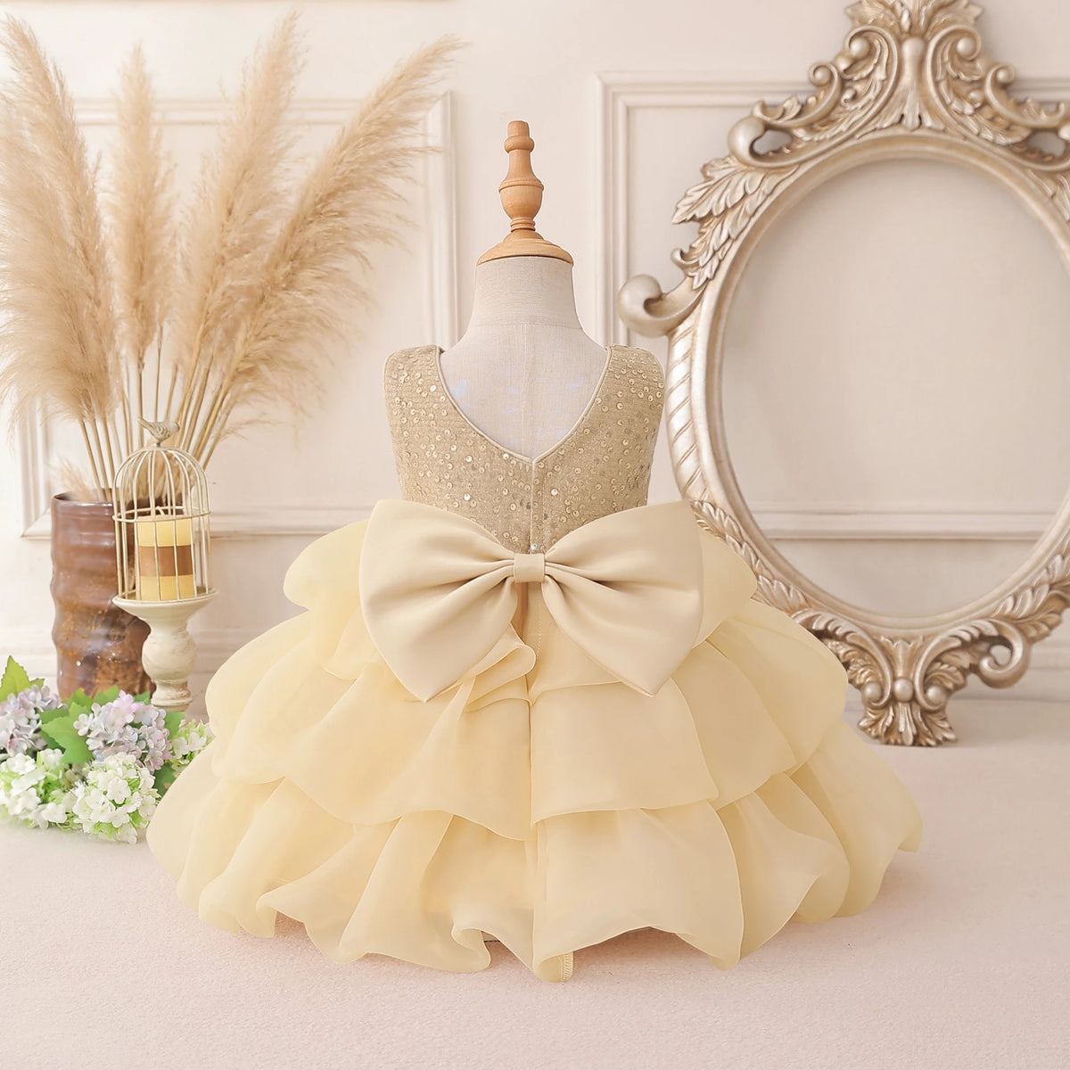 A toddler girl's formal party dress displayed on a mannequin, featuring a sparkly gold sequined bodice, a large cream-colored ruffled bow at the waist, and a tiered ruffled skirt. The dress is styled in a vintage-inspired room setting with a large ornate mirror, pampas grass, and flowers.