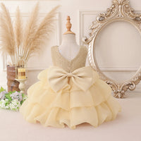 A toddler girl's formal party dress displayed on a mannequin, featuring a sparkly gold sequined bodice, a large cream-colored ruffled bow at the waist, and a tiered ruffled skirt. The dress is styled in a vintage-inspired room setting with a large ornate mirror, pampas grass, and flowers.
