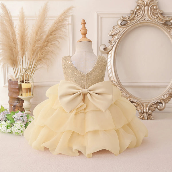 A toddler girl's formal party dress displayed on a mannequin, featuring a sparkly gold sequined bodice, a large cream-colored ruffled bow at the waist, and a tiered ruffled skirt. The dress is styled in a vintage-inspired room setting with a large ornate mirror, pampas grass, and flowers.