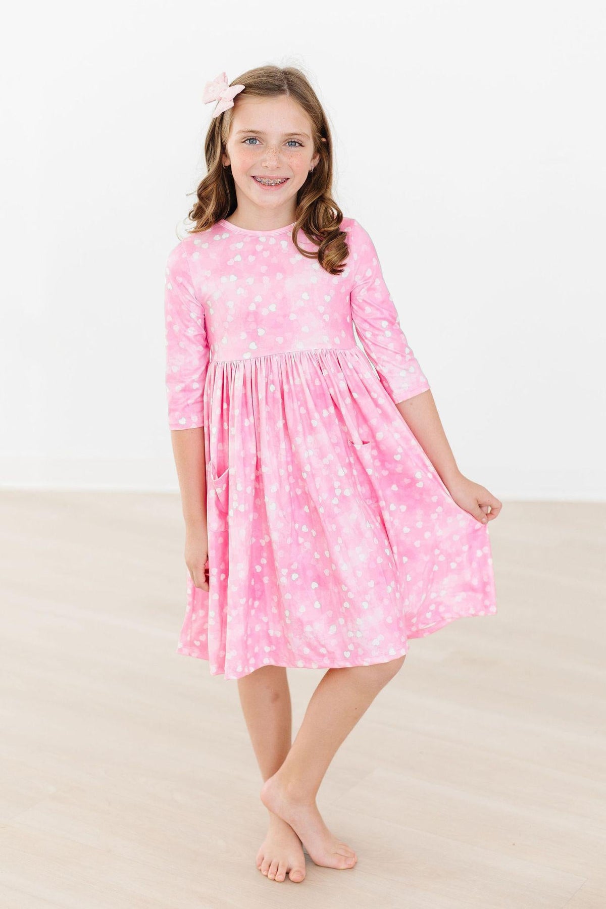 Girl wearing pink 3/4 sleeve twirl dress with white heart print, scoop neck, and side pockets for play.